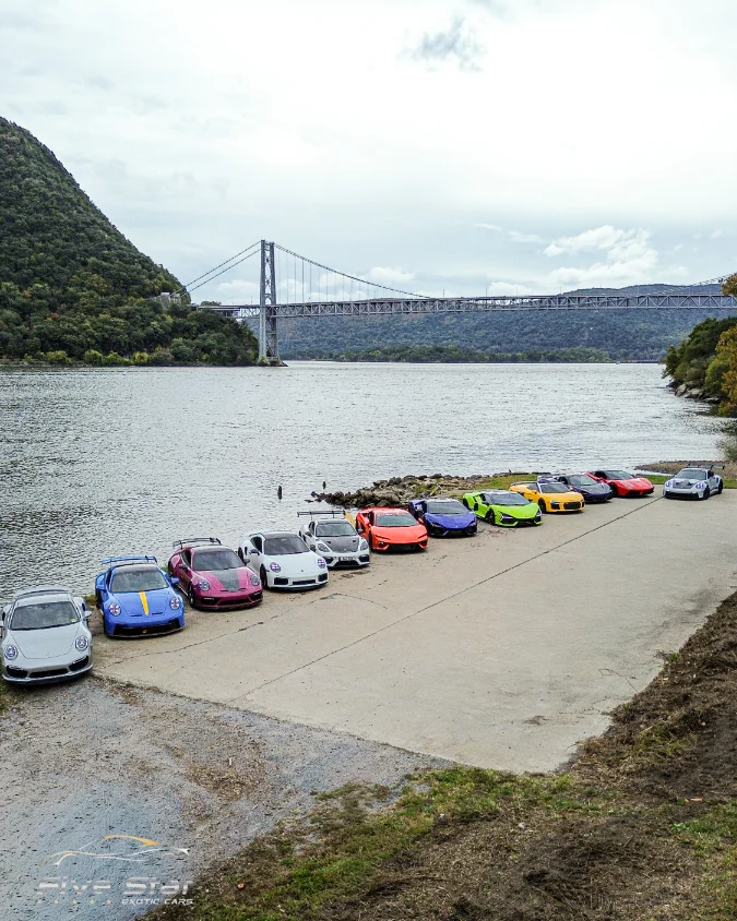 A line of colorful sports cars beside the river, with a suspension bridge beyond.