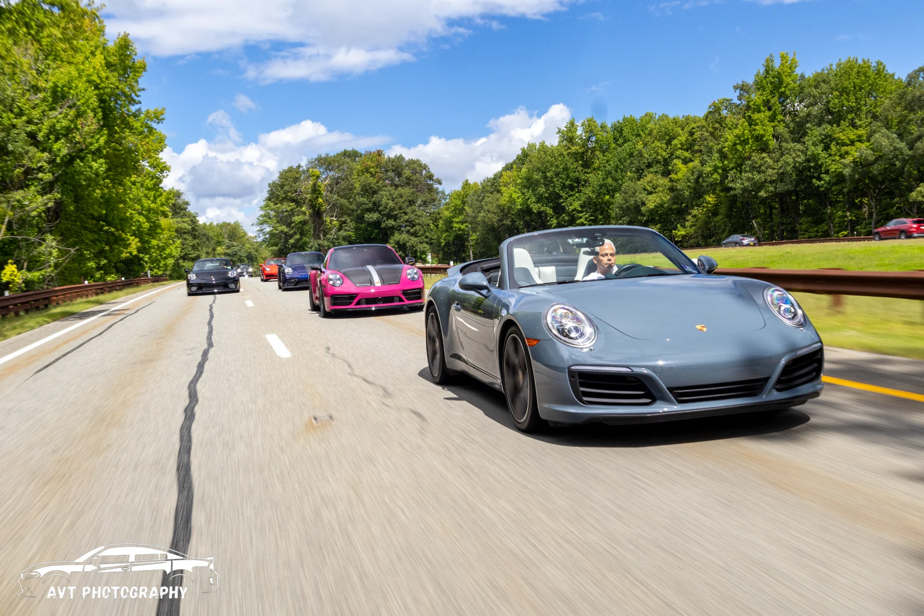 A silver sports car and a convoy travelling along a leafy road.